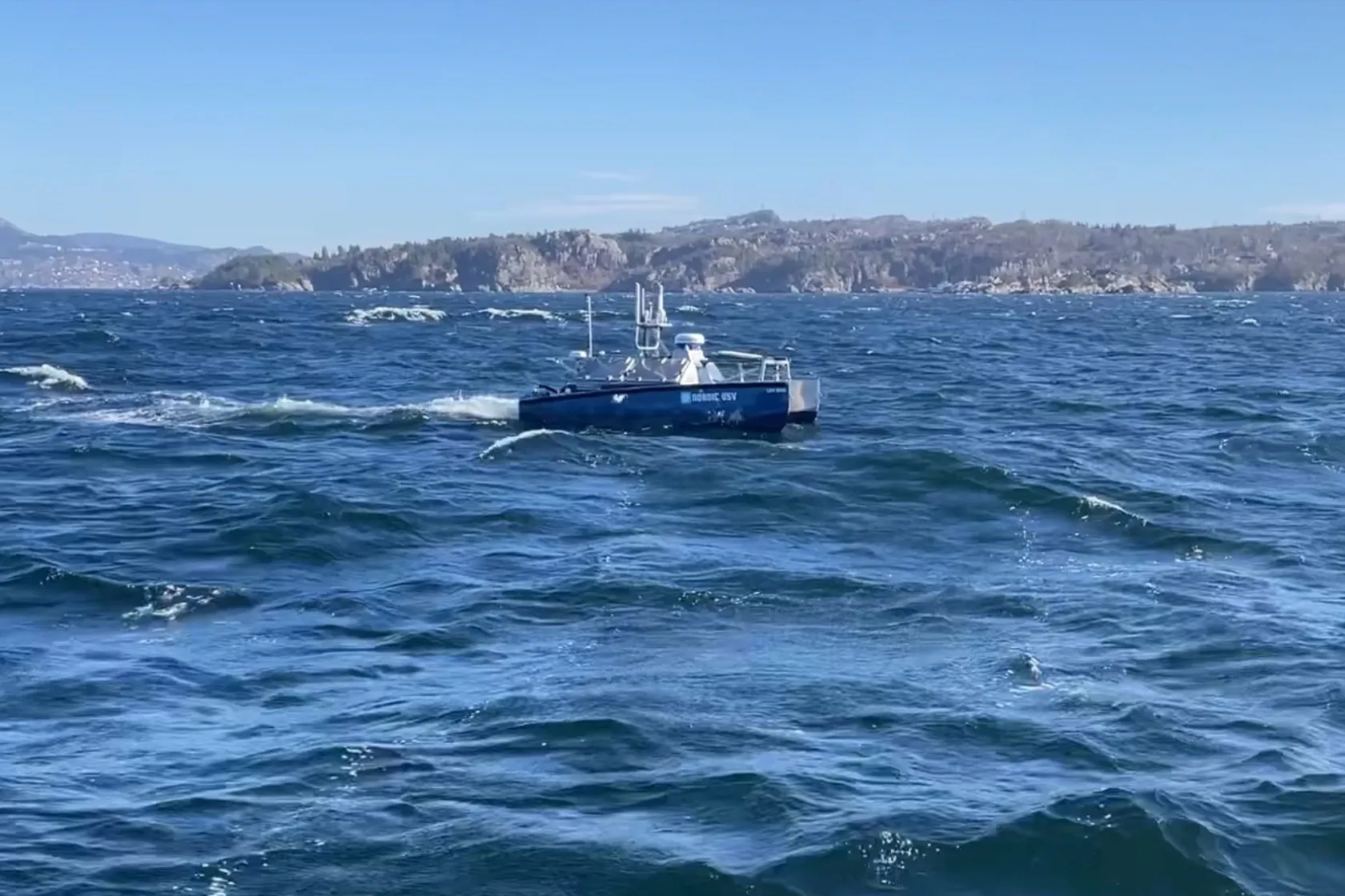 Ubemannet båt fra Nordic USV i krapp sjø med skumtopper, med kysten i bakgrunnen.